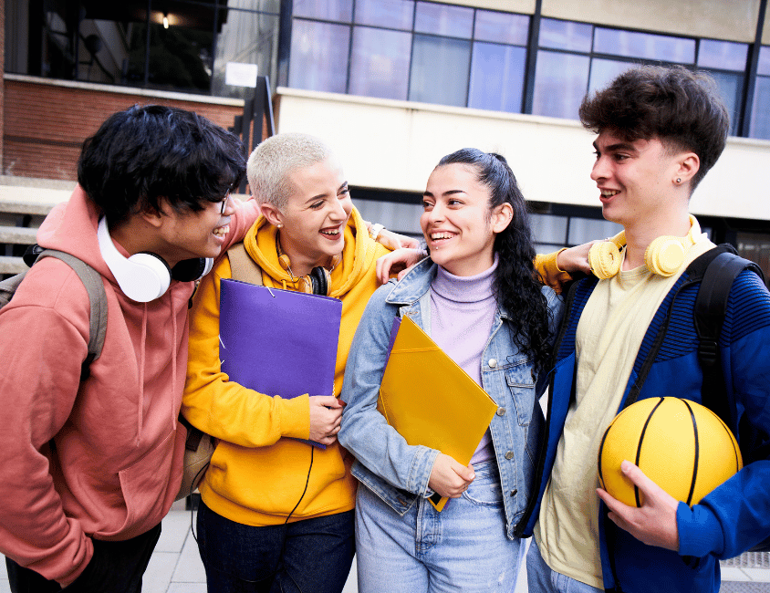 A group of young individuals, diverse in appearance and style, are gathered outside an apartment building. They are engaged in a lively and enjoyable conversation, with smiles on their faces, as they share stories and connect with one another on a beautiful day