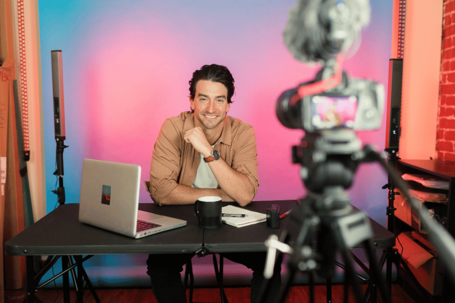 An influencer, set up with a tripod, laptop, lighting, and camera, smiles at the camera, ready to film his next video.