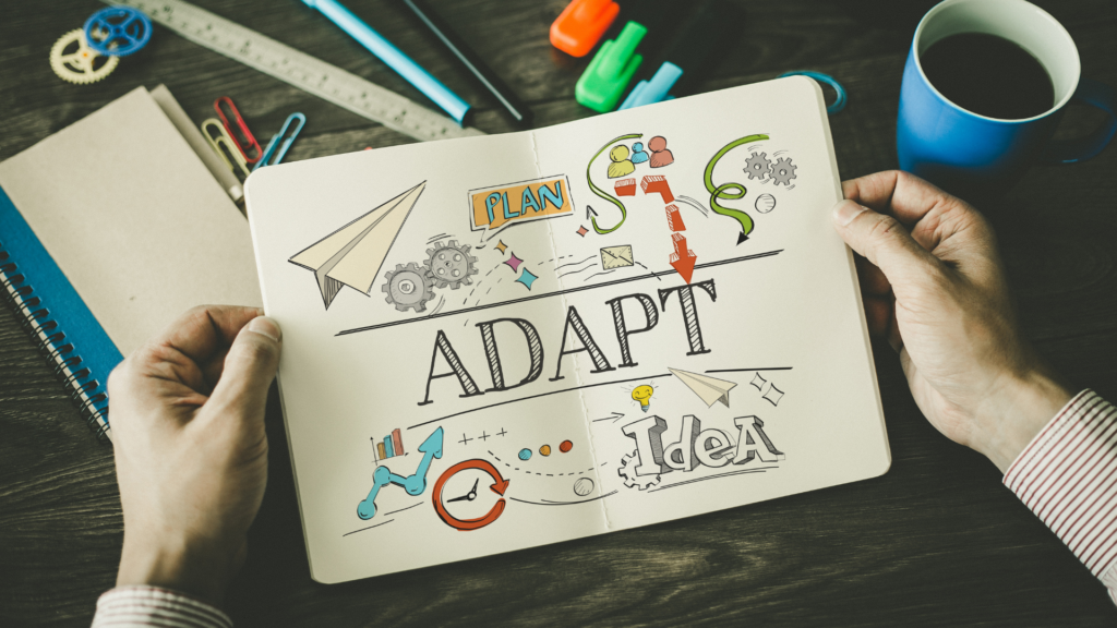 Hands holding a notebook opened to a page with the word “adapt” written on it surround by office supplies.