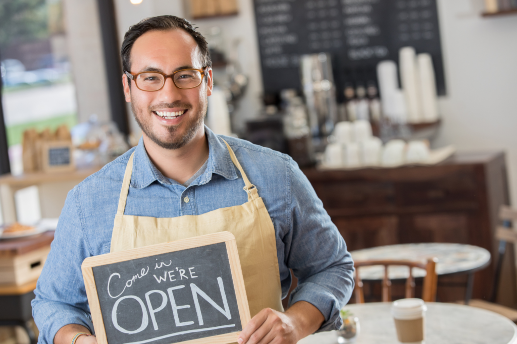 Man standing outside his small business coffee shop holding a "Come In, We're Open" sign, representing marketing for small business through strong local branding and customer engagement.