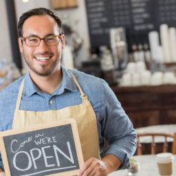 Man standing outside his small business coffee shop holding a "Come In, We're Open" sign, representing marketing for small business through strong local branding and customer engagement.